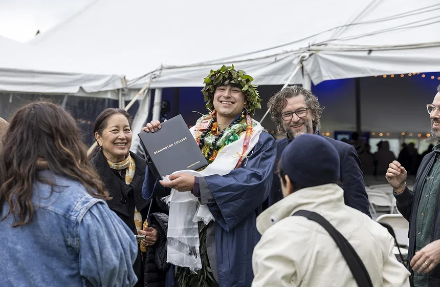 A male Hawaiian student, wearing a traditional head dress and lei, stands looking at the camera and holding up his diploma while surrounded by his family with his family 