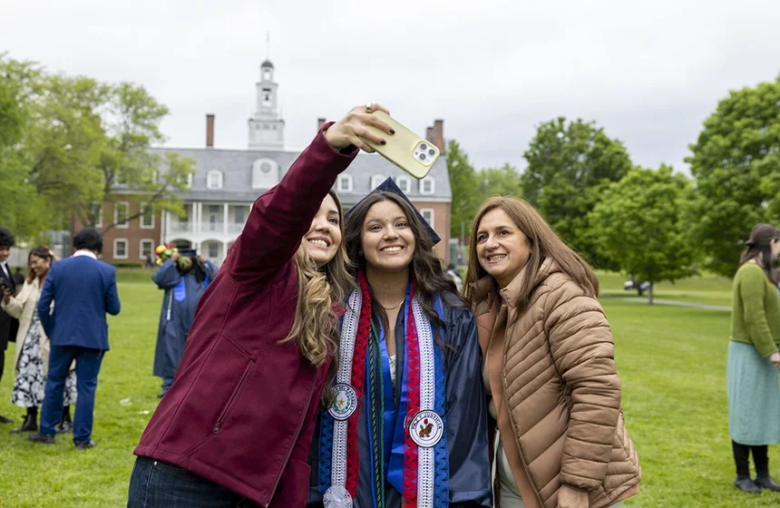 A family, with their back to commons, student in cap and gown at the center, take a selfie