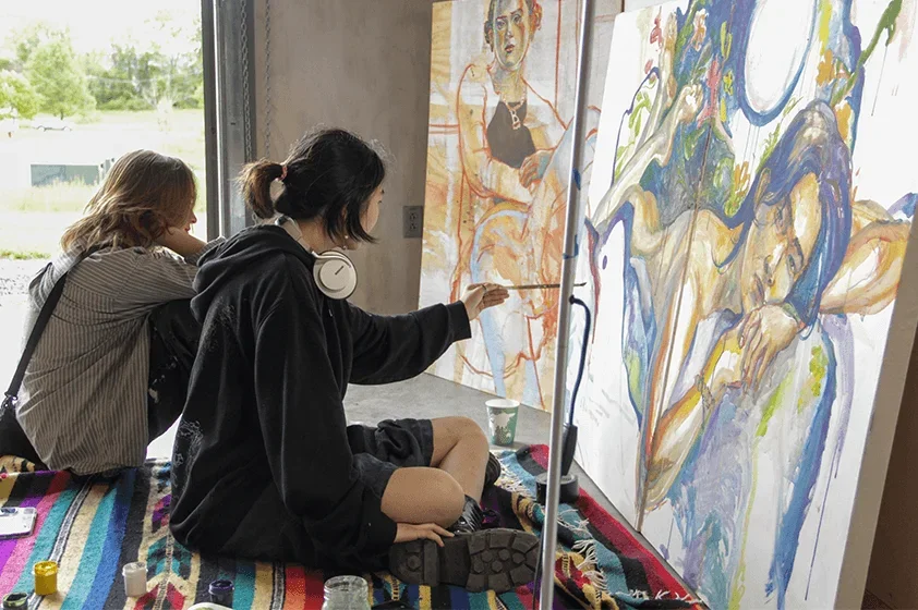 two students sit beside each other while they actively work on their paintings
