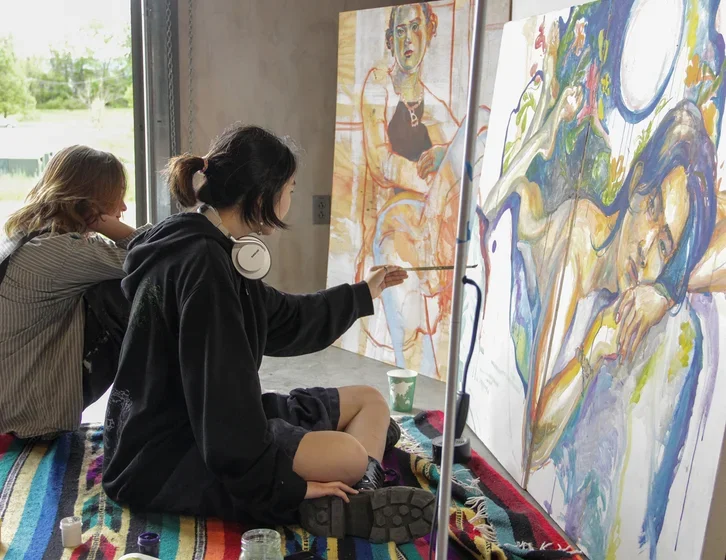 student wearing a black hooded sweatshirt sitting cross-legged on the floor and painting a figure on a large horizontal canvas